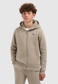 Jonge man in een beige rits-hoodie en bijpassende sweatpants, staand met één hand in de zak tegen een effen achtergrond.