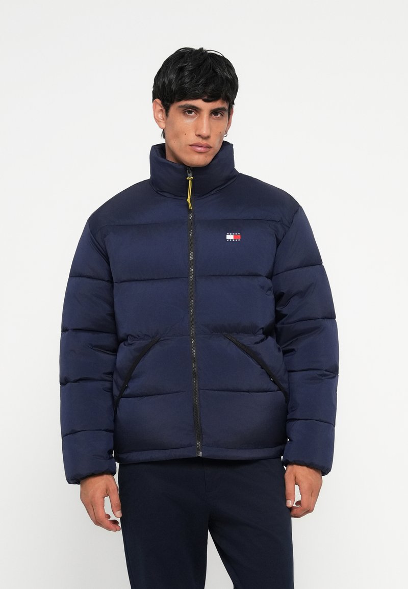 Marineblauer Pufferjacke mit hohem Kragen, ausgestattet mit einem gelben Reißverschluss. Mit schwarzem Besatz und Fronttaschen versehen, bietet sie ein gepolstertes Design.