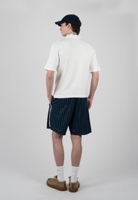 Jeune homme vu de dos portant une chemise blanche à manches courtes, un short rayé bleu marine, des chaussettes blanches, des chaussures marron et une casquette de baseball bleu marine.