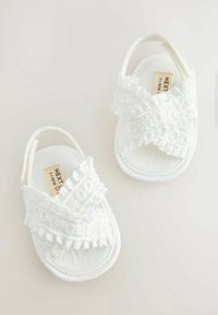 Bílé dětské sandály s texturovaným designem křížového pásku, zdobené volánky. Vyrobené z měkké látky s plochou podrážkou pro pohodlí.