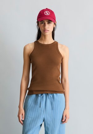 Młoda kobieta w czerwonej czapce, brązowym bezrękawniku żebrowanym i jasnoniebieskich spodniach w cienkie paski z sznurkiem, na jednolitym szarym tle.