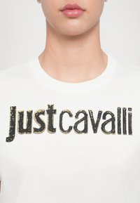 Valge puuvillane T-särk ümmarguse kaelusega, mille esiküljel on suur, kulunud must ja kuldne "Justcavalli" logo.