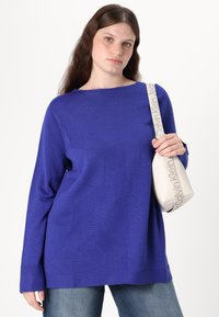 Pulover albastru tricotat cu un decolteu larg și mâneci lungi, având un model texturat. Modelul ține o geantă de culoare crem cu un braț.