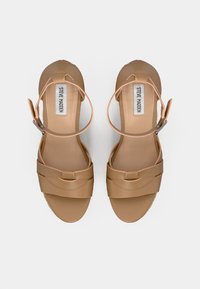 Sandales strappy beiges avec une sangle au niveau de la cheville, fabriquées en matériau synthétique lisse, présentant des bandes supérieures croisées et une semelle plate. Accents en équipement argenté.