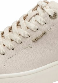 Približan posnetek bež usnjenega športnega copata z debelimi kremnimi vezalkami, kovinskimi zankami in logotipom blagovne znamke "Tamaris" na strani.