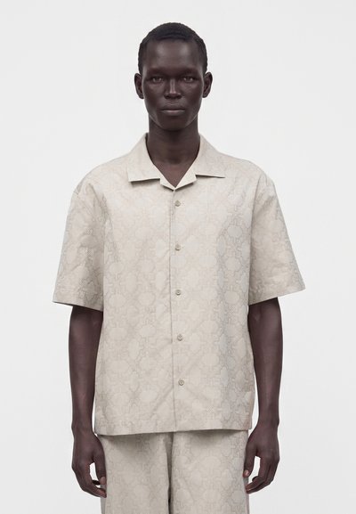 Homme portant une chemise beige clair à manches courtes boutonnée et un short assorti avec un motif géométrique subtil, debout devant un fond uni.