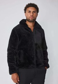 Homme portant une veste zippée noire en peluche avec poche poitrine zippée, se tenant contre un fond gris clair uni.