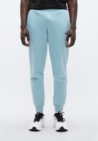 Ανοιχτό μπλε φόρμα παντελόνι με ελαστική μέση και μανσέτες, κατασκευασμένο από απαλό υλικό. Διαθέτει λογότυπο PUMA στο αριστερό μηρό. Συνδυάζεται με μαύρα και λευκά αθλητικά παπούτσια.