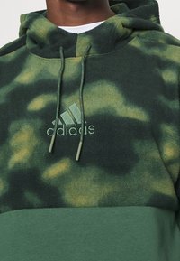Felpa verde con camouflage realizzata in materiale fleece, dotata di cappuccio regolabile con coulisse, caratterizzata da una finitura testurizzata e un logo Adidas ricamato nello stesso colore.