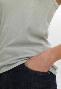 Heled hall ribaga tank top, mis on valmistatud tekstureeritud kangast, sobib tumedate siniste denimiste teksadega. Käsi puhkab teksadest taskus.