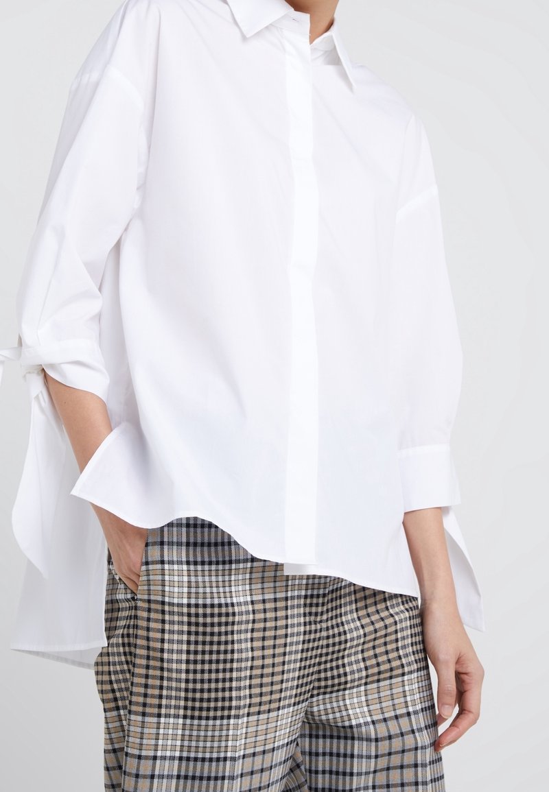 Blouse en coton blanc avec fermeture à boutons, coupe oversize et détails plissés ; associée à un pantalon à motif vichy dans des tons beige et bleu marine.
