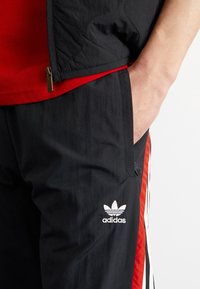 Svarta träningsbyxor med röda detaljer och vit Adidas-logotyp. Har en slät textur, sidofickor och kontrasterande ränder längs sidorna.