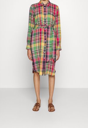 Femme portant une robe chemise à carreaux colorés avec manches longues, boutonnage devant, ceinture à la taille et sandales plates marron sur un fond uni.