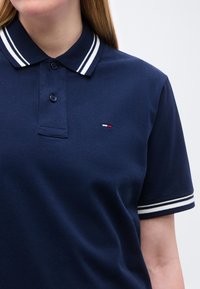 Femeie purtând un tricou polo albastru închis cu guler și manșete cu dungi albe, având un mic logo roșu, alb și albastru pe piept.