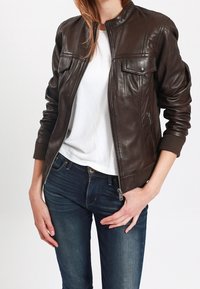 Brązowa skórzana kurtka bombowiec z zamkiem błyskawicznym, dwoma kieszeniami na piersi i ściągaczami na rękawach. Nosi się ją na białej koszuli i ciemnoniebieskich dżinsach.