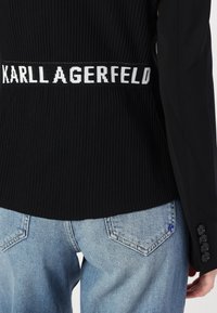 Černý svetřík z ribbené látky s logo páskem s nápisem "KARL LAGERFELD" a manžetami na jeden knoflík, v kombinaci se světle modrými džíny.