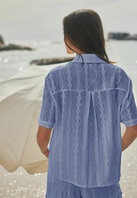 Donna con capelli lunghi e scuri che indossa una camicia azzurra a fantasia e pantaloncini, in piedi vicino al mare con un ombrellone da spiaggia beige sullo sfondo.