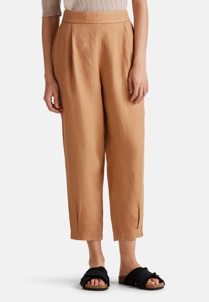Pantalones de lino color marrón claro con cintura alta, pliegues frontales y puños a la altura del tobillo. Llevados con sandalias de diapositivas negras y esponjosas.