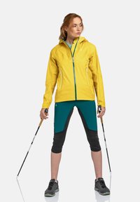 Geel waterdichte jas met een capuchon, voorzien van een rits aan de voorkant en twee zijzakken. Geko combineerd met teal shorts en zwarte legging, terwijl hij trekkingstokken vasthoudt.