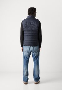 Colete de puffer acolchoado azul-marinho com gola alta, usado sobre uma camisa preta de mangas longas, combinado com jeans azul desbotado e calçado castanho.