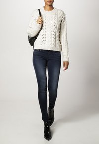 Biały sweter z dzianiny warkoczowej z fakturowanymi wzorami, zestawiony z ciemnoniebieskimi obcisłymi dżinsami i czarnymi botkami. Modelka trzyma czarną torebkę.