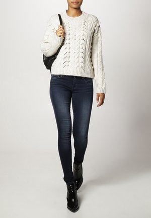 Witte kabelgebreide trui met textuurpatronen, gecombineerd met donkerblauwe skinny jeans en zwarte enkellaarsjes. Model houdt een zwarte tas vast.