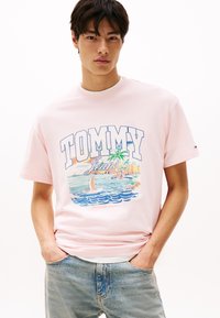 Rožinė medvilnės marškinė su dideliu „TOMMY Jeans“ grafiniu piešiniu, vaizduojančiu paplūdimio sceną su jachtomis ir palmėmis. Su apvalia iškirpte.
