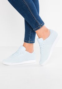 Personne portant des baskets en daim bleu clair avec des semelles blanches et un jean bleu ajusté se tenant sur un sol blanc.