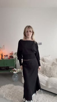 Γυναίκα με ξανθά μαλλιά που φοράει μαύρη μπλούζα μακρυμάνικη και ταιριαστή φούστα, στέκεται σε ένα ζεστό σαλόνι με ανοιχτή διακόσμηση.