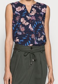 Blouse sans manches florale marine avec un col en V, présentant des motifs roses et bleus sur un tissu texturé, associée à un pantalon vert taille haute.