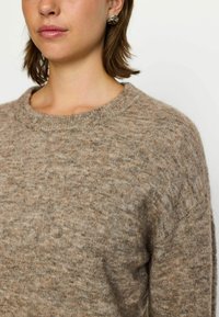 Pull beige en tricot avec un col rond. Texture douce, finition légèrement duveteuse, coupe ample et detailing minimal sur les manches.