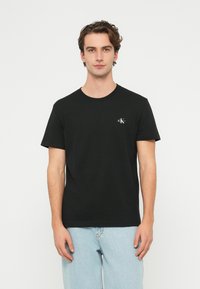 Fekete pamut póló kerek nyakkivágással, rövid ujjú, kis fehér logóval a mellkason. Világoskék farmernadrággal viselve.