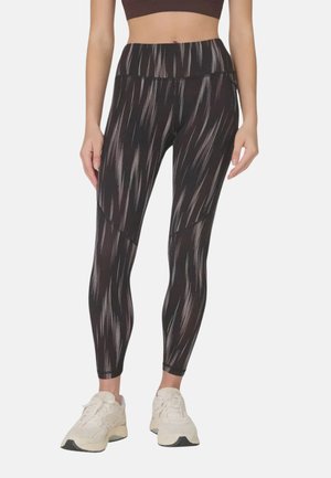 Vrouw die zwart-wit patroonleggings met hoge taille en witte sportschoenen draagt, staand met ontspannen armen langs haar zij.