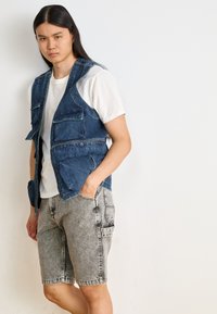 Jeans prsluk s višestrukim džepovima, natrag preko bijele majice. Uparen sa sivim, izlizanim šortsevima. Opuštena kroja i utilitarni dizajn.