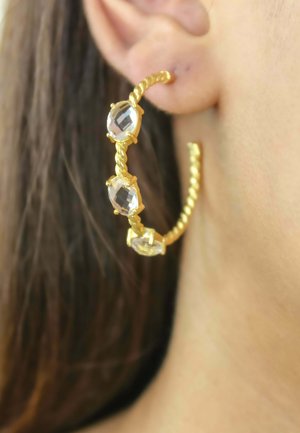 Boucle d’oreille anneau torsadé en or avec trois pierres précieuses facettées claires portée à l'oreille gauche avec des cheveux foncés en arrière-plan.