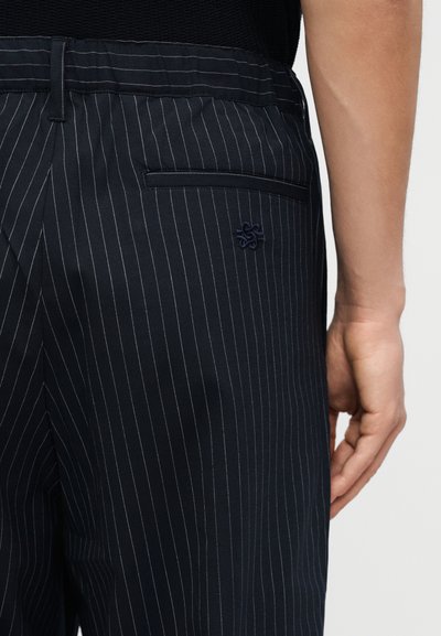 Κοντινό πλάνο ενός ατόμου που φοράει σκούρα μπλε παντελόνια με ρίγες, πίσω τσέπη με φάσα και κεντημένο λογότυπο κοντά στην τσέπη.