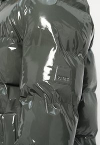 Jachetă căptușită din material lucios de culoare gri închis, cu o textură netedă. Prezintă o patch cu logo pătrat pe mânecă și un design pufos.