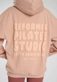 Rosa huvtröja med plysch, överdimensionerad text: "REFORMER PILATES STUDIO SOUTH BEACH MIAMI GRUNDAT 1953" i en något ljusare nyans.