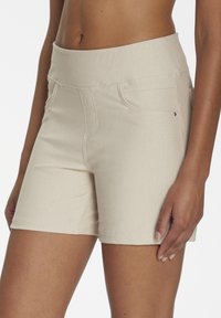 Close-up of a person wearing beige fitted shorts with front pockets, showing midriff and part of the right arm and hand.