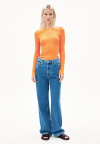 Rippiges Langarmoberteil in leuchtendem Orange, kombiniert mit hellblauen, weiten Jeans. Das Modell trägt dunkle Slippers. Einfaches, klares Design.