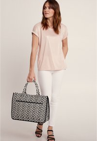 Helles Rosa, kurzärmeliges Oberteil kombiniert mit weißen Hosen. Das Modell trägt eine schwarz-weiß gemusterte Tasche mit zwei Griffen und einem Reißverschluss.