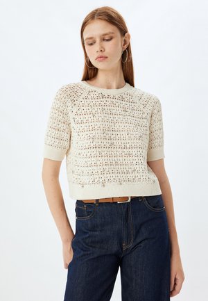 Crème gebreide trui met korte mouwen, opengewerkt patroon en geribde zoom. Gecombineerd met donkerblauwe denim broek, wat de textuurcontrast benadrukt.