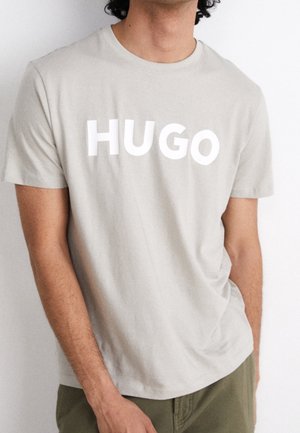 Man som bär en ljusbeige T-shirt med "HUGO" tryckt i stora vita bokstäver, tillsammans med olivgröna byxor.