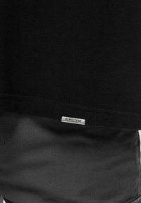 Camiseta de algodón negra con dobladillo recto, con una etiqueta plateada "REPRESENT" en la parte inferior. Combinada con pantalones negros texturizados.