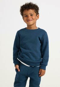 Felpa in cotone blu con colletto tondo e polsini a coste, indossata sopra una camicia bianca, abbinata a pantaloni blu coordinati. Bambino che sorride.