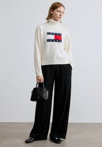 Pull en maille blanche à col roulé avec logo brodé "TOMMY JEANS", associé à un pantalon noir à jambes larges et un petit sac à main noir.