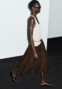 Modello che indossa un top beige, una gonna fluida marrone e sandali neri. Gli accessori includono occhiali da sole scuri e una cintura con fibbia circolare.