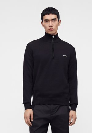 Modèle masculin portant un pull noir zippé avec le logo "HUGO" sur la poitrine et un pantalon noir, debout devant un fond clair uni.