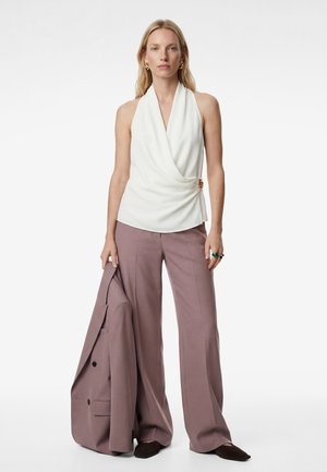 Frau trägt weißes ärmelloses, drapiertes Oberteil mit goldener Brosche, mauvefarbene Hosen mit weitem Bein, hält passenden Blazer und trägt goldene Ohrringe.