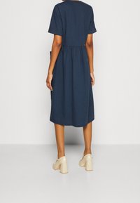 Kvinna iklädd en marinblå midiklänning och beige klackskor står mot en enhetlig bakgrund, sett bakifrån.